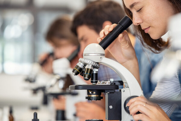 Three students in white lab coats looking into microscopes on a lab bench, with glassware and equipment visible behind them.