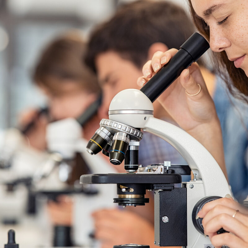 Three students in white lab coats looking into microscopes on a lab bench, with glassware and equipment visible behind them.