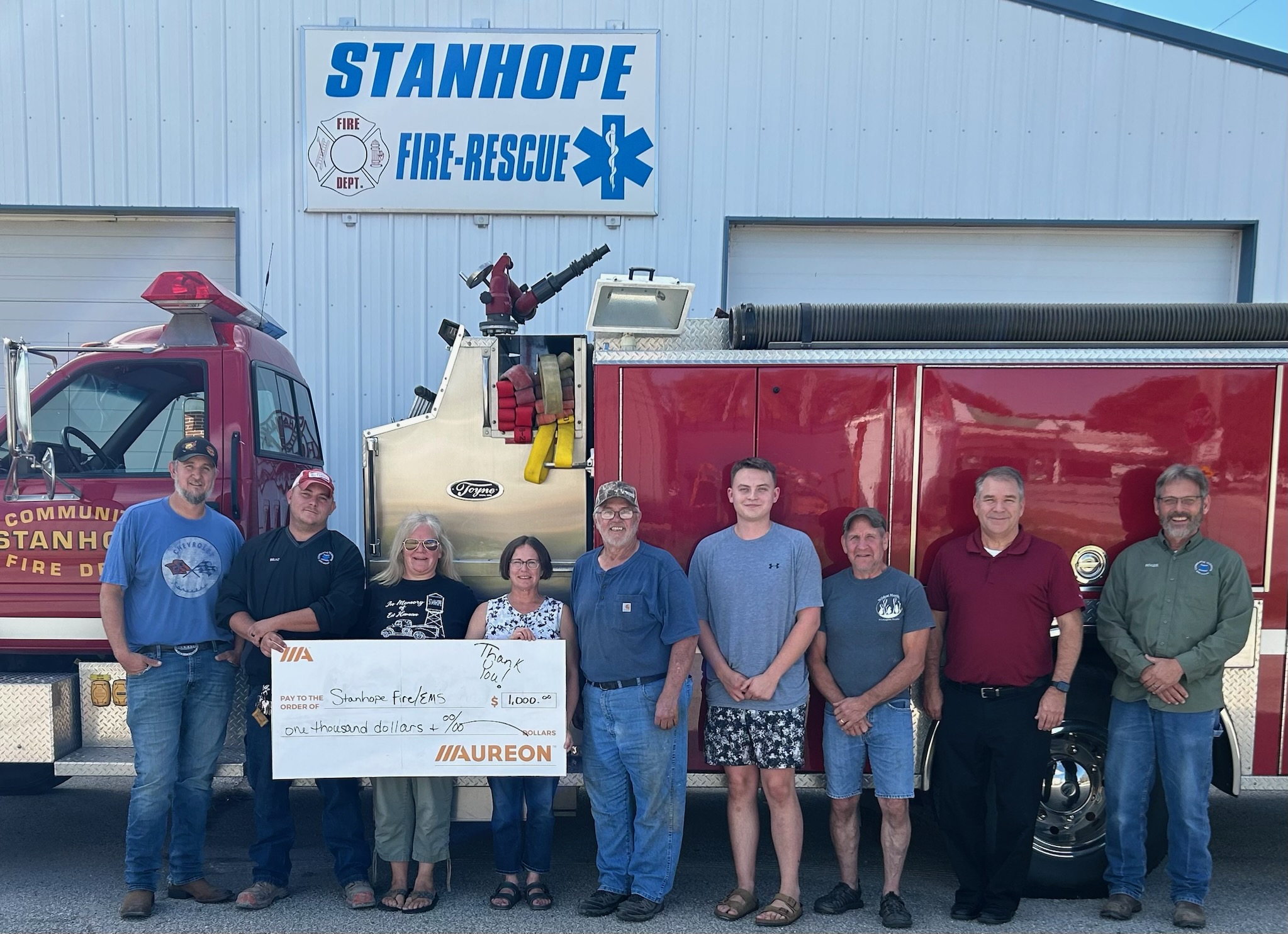 Nine Stanhope Fire-Rescue personnel standing in front of a fire truck and station sign, holding a giant Aureon check for $1,000.”