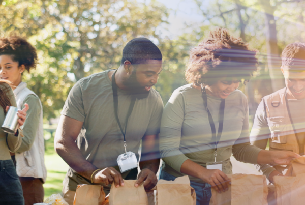 Five volunteers in a park filling brown paper bags with food items at an outdoor table under trees.
