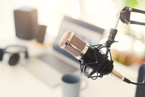 Close-up of a condenser microphone on a boom arm with a blurred laptop, headphones, and coffee mug on a desk in the background.