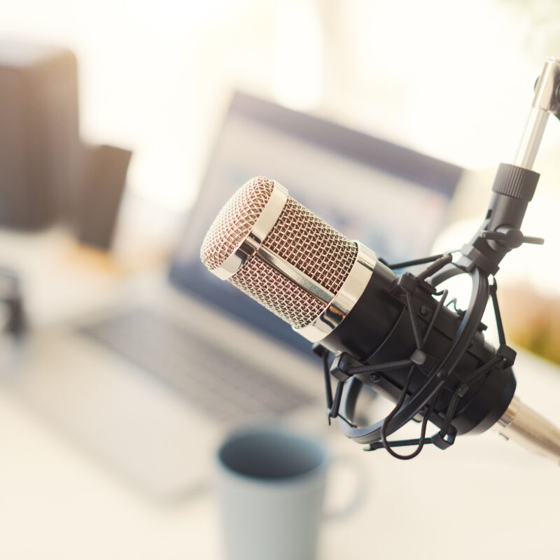 Close-up of a condenser microphone on a boom arm with a blurred laptop, headphones, and coffee mug on a desk in the background.