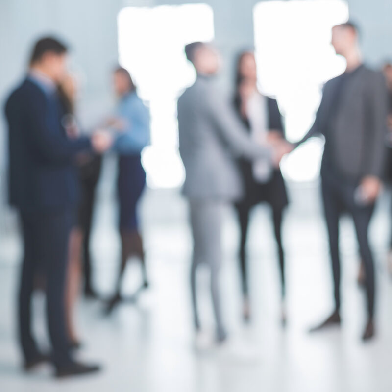 Blurred group of business professionals in suits milling about and two people shaking hands in a bright modern office lobby.