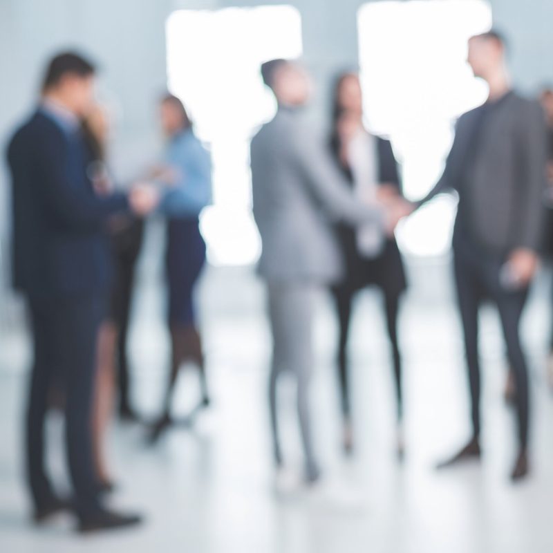 Blurred group of business professionals in suits milling about and two people shaking hands in a bright modern office lobby.