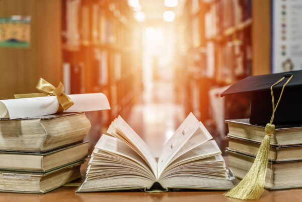 An open old textbook on a wooden desk surrounded by stacks of books, with blurred library shelves in the background.”