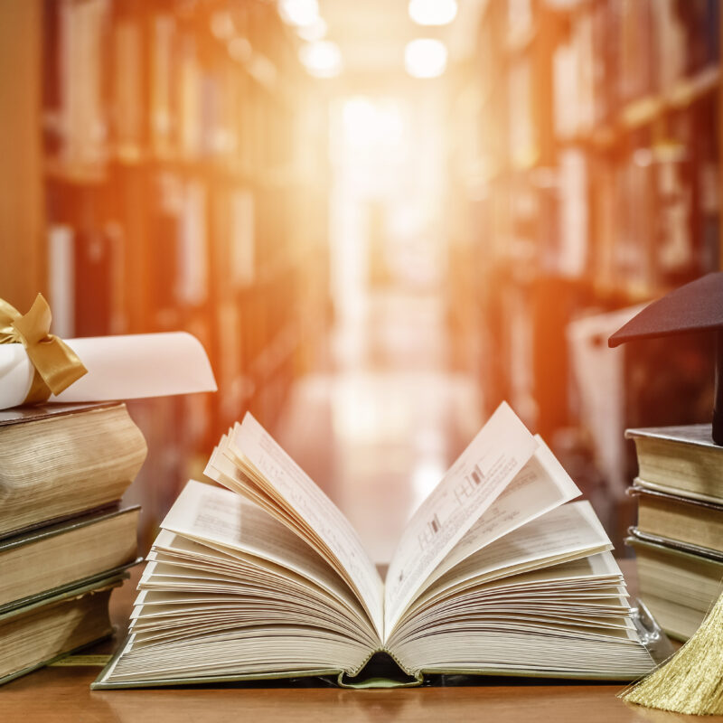 An open old textbook on a wooden desk surrounded by stacks of books, with blurred library shelves in the background.”