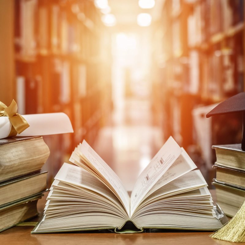 An open old textbook on a wooden desk surrounded by stacks of books, with blurred library shelves in the background.”