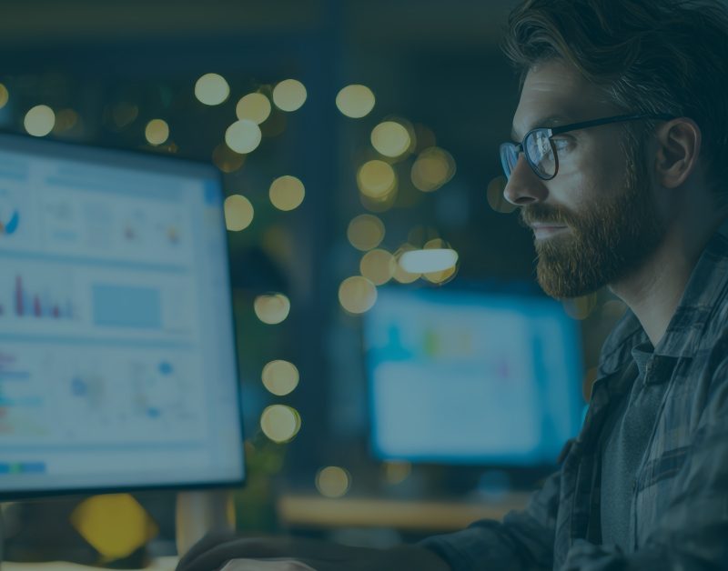 Professional reviewing Security Awareness Training analytics dashboard on dual monitors, analyzing phishing simulation results and employee security performance data in a low-light office setting.