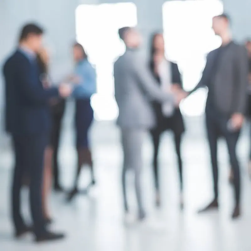 Blurred group of business professionals in suits milling about and two people shaking hands in a bright modern office lobby.