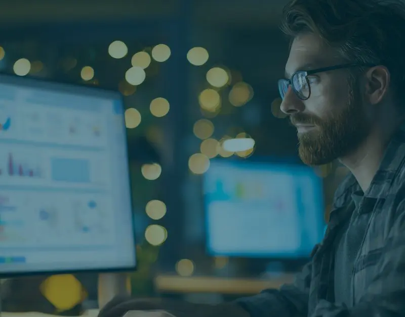 Professional reviewing Security Awareness Training analytics dashboard on dual monitors, analyzing phishing simulation results and employee security performance data in a low-light office setting.