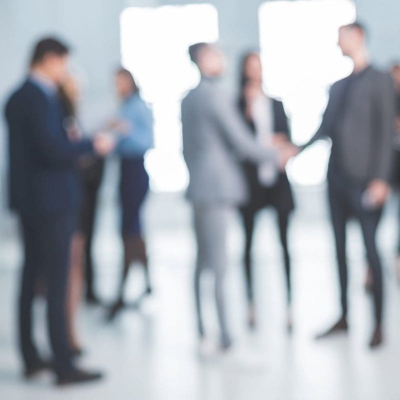 Blurred group of business professionals in suits milling about and two people shaking hands in a bright modern office lobby.