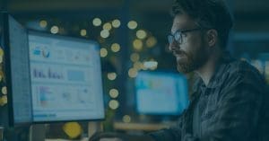 Professional reviewing Security Awareness Training analytics dashboard on dual monitors, analyzing phishing simulation results and employee security performance data in a low-light office setting.